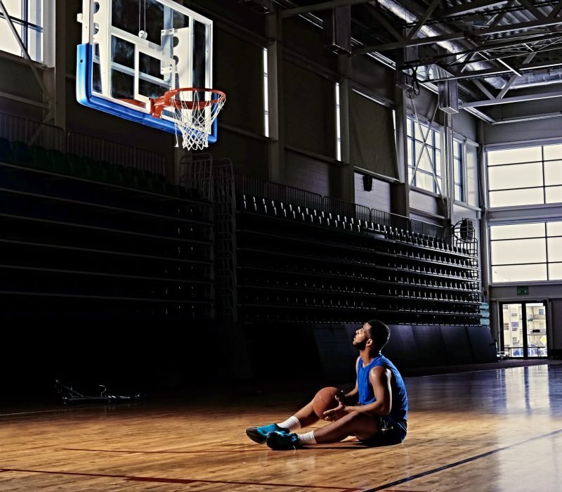 Black professional basketball player sits on a field under the hoop in a game hall.
